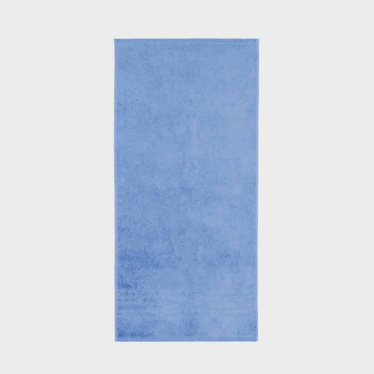 A simple light blue BRILLIANT towel (600 g/m²) lies flat on a white background. The towel has a soft, plush texture and a slightly darker blue band runs along one end.